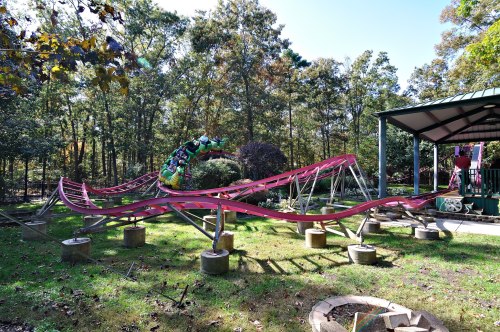 A colorful roller coaster in a wooded area, with tracks curving and a small shelter nearby.