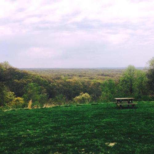 A scenic view of rolling hills and trees under a cloudy sky, with a picnic table in the foreground.