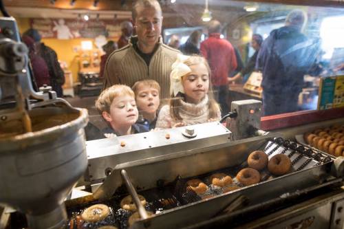 A man and three children watch donuts being made at a shop, with warm lighting and a busy atmosphere.