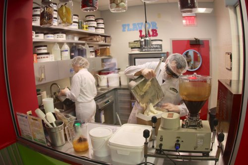 Two people in lab coats work in a colorful kitchen, mixing ingredients in a flavor lab. Containers and equipment are visible.