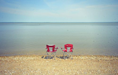 Two red folding chairs sit on a sandy beach by a calm lake, with a clear blue sky above.
