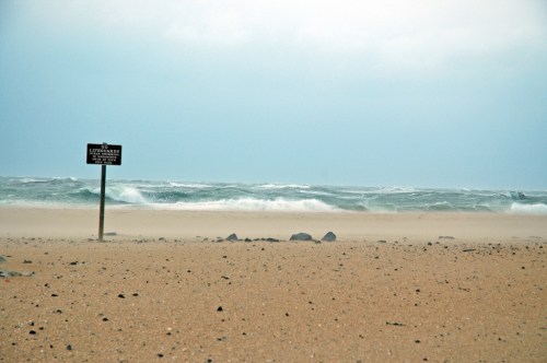 A sandy beach with a signpost and turbulent waves in the background under a cloudy sky.