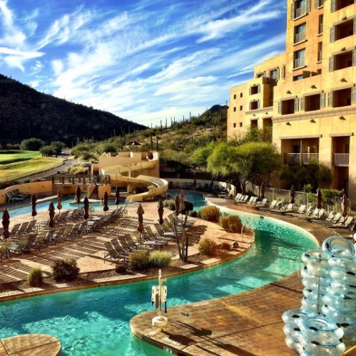 A scenic hotel pool area with lounge chairs, umbrellas, and mountains in the background under a blue sky.
