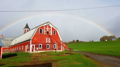 A red barn stands in a green field under a bright rainbow against a cloudy sky.