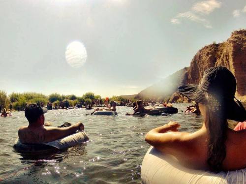 A crowded river scene with people floating on inner tubes, surrounded by cliffs and greenery under a sunny sky.