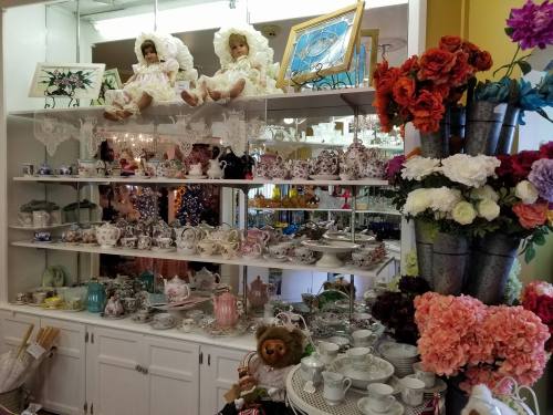 A display of vintage teapots, porcelain dishes, and decorative dolls, surrounded by colorful flowers.