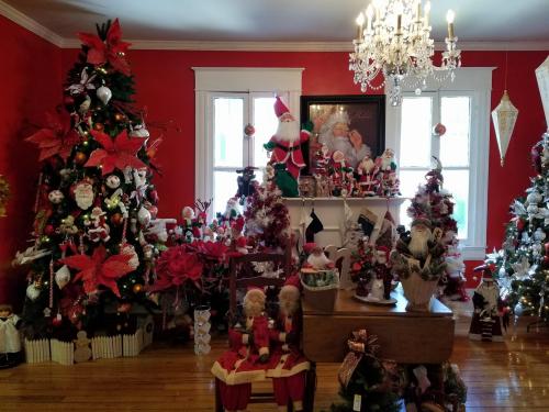 A festive room filled with decorated Christmas trees, ornaments, and holiday decorations, featuring a chandelier above.