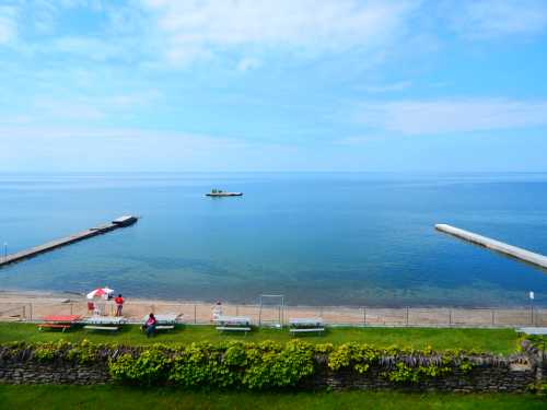 A calm lake scene with a sandy beach, docks, and a boat in the distance under a blue sky with scattered clouds.