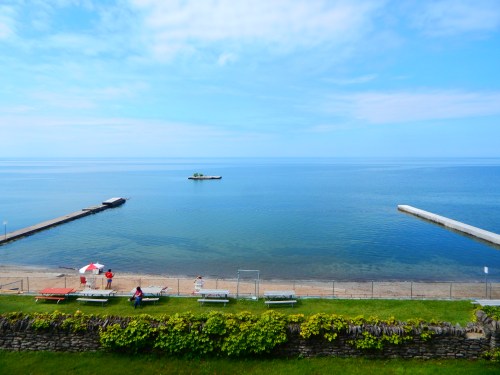 A calm lake scene with a sandy beach, docks, and a boat in the distance under a blue sky with scattered clouds.