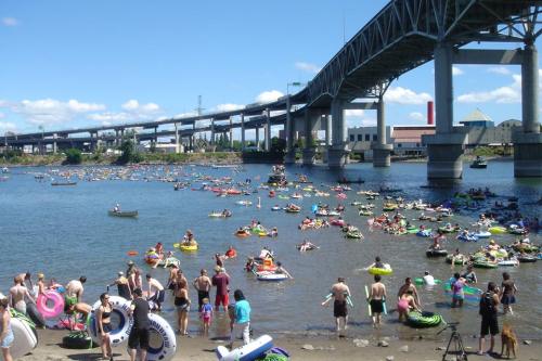 A crowded river scene with people floating on inner tubes and enjoying the sun under a bridge.