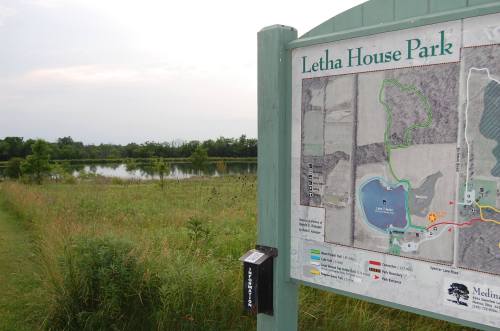 Sign for Letha House Park with a map, surrounded by grassy areas and a calm water body in the background.