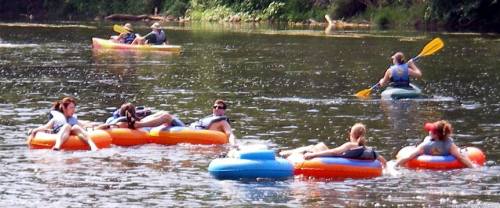 A group of people floating on colorful tubes in a river, with a kayaker paddling nearby. Lush greenery surrounds the scene.