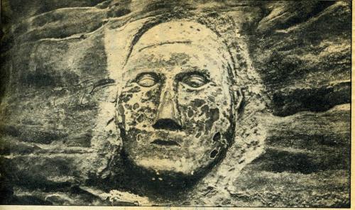 A weathered stone carving of a human face, partially eroded, set against a rocky background.