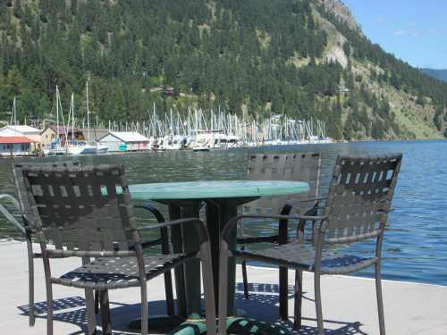 A table and chairs on a dock overlooking a calm lake with boats and a forested hillside in the background.