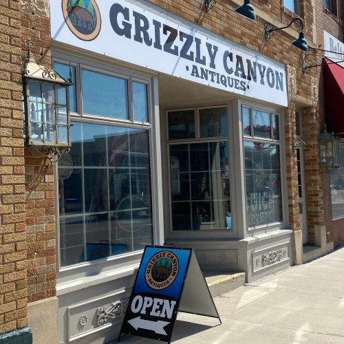 Exterior of Grizzly Canyon Antiques store with an "Open" sign and large windows on a sunny day.