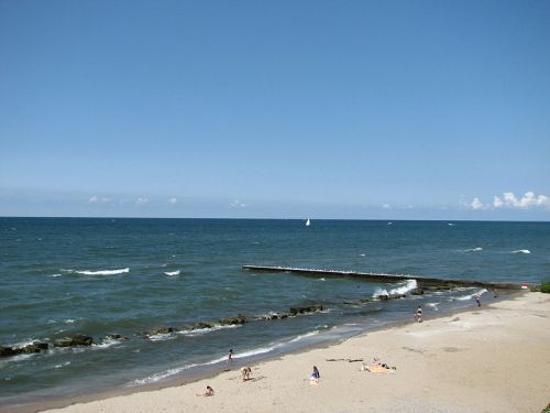 A sunny beach scene with people relaxing on the sand and a sailboat in the distance on a calm sea.