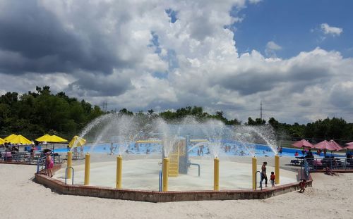 A splash pad with water fountains, surrounded by children playing and colorful umbrellas on a sunny day.