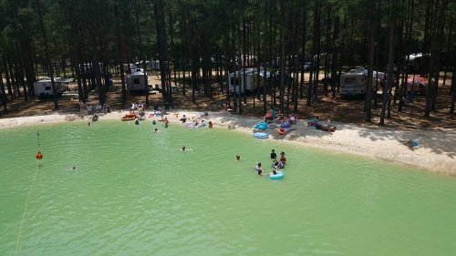 A serene lake surrounded by trees, with people swimming and relaxing on inflatable floats. RVs are visible in the background.