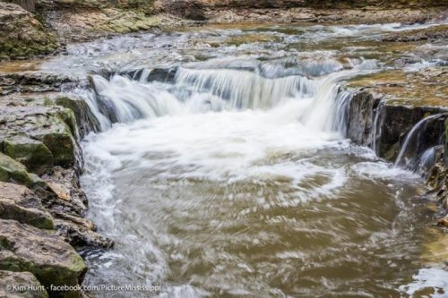 A serene waterfall cascades over rocky terrain, creating ripples in a tranquil stream.