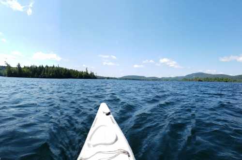 A kayak's bow cutting through calm water, surrounded by lush green trees and distant mountains under a clear blue sky.