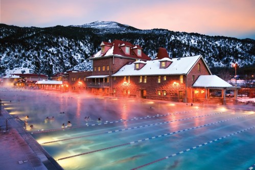 A scenic outdoor hot spring pool surrounded by snow-covered mountains at sunset, with steam rising and people enjoying the water.