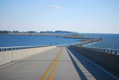 A wide road leads towards a bridge over water, with an island visible in the distance under a clear blue sky.