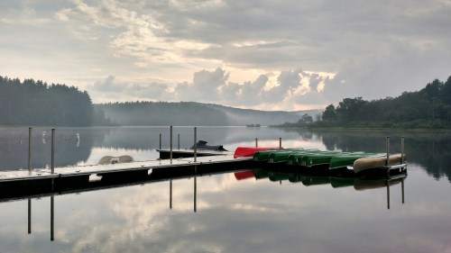 A calm lake at dawn with a wooden dock and colorful boats, surrounded by misty trees and soft clouds.