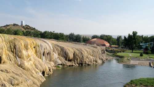 A scenic view of a river flowing past a rocky, mineral-laden cliff with greenery and a building in the background.