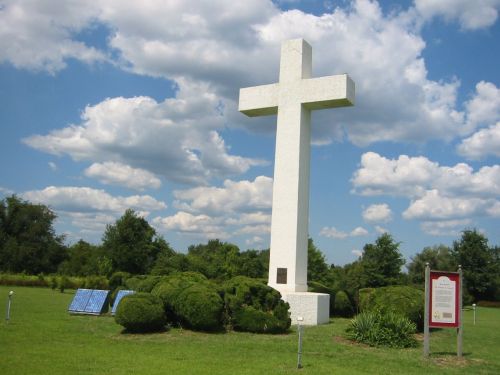A large white cross stands in a grassy area, surrounded by bushes and under a blue sky with fluffy clouds.