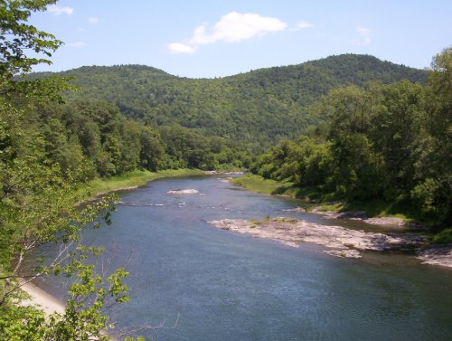 A serene river flows through lush green hills under a clear blue sky, with rocky banks and scattered trees.