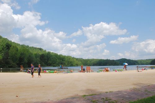 A sandy beach by a lake, with people walking, lounging, and enjoying the sunny day under a partly cloudy sky.