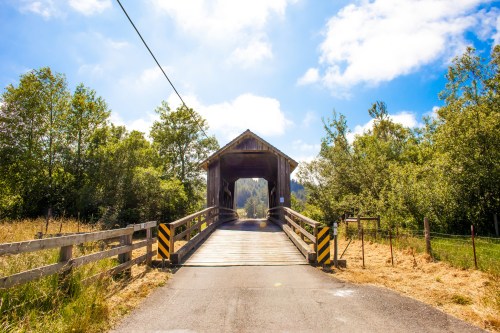 A wooden covered bridge spans a road, surrounded by lush greenery and a bright blue sky with scattered clouds.