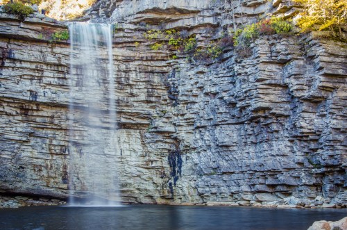 A serene waterfall cascading down rocky cliffs into a calm pool below, surrounded by autumn foliage.
