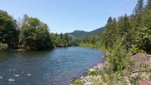 A serene river flows through a lush green landscape, surrounded by trees and mountains under a clear blue sky.