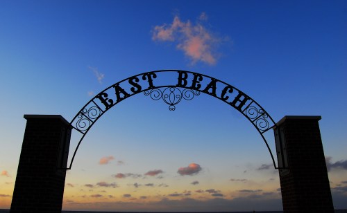 Silhouette of an archway sign reading "East Beach" against a colorful sunset sky.