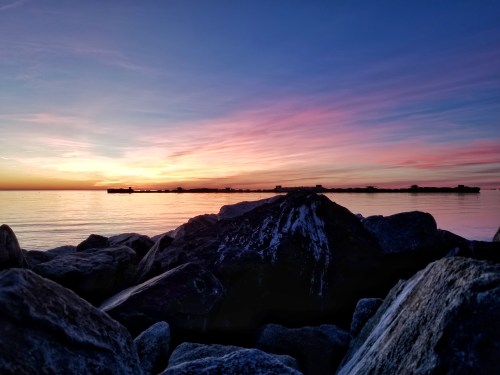 A serene sunset over calm waters, with colorful clouds reflecting on the surface and rocky foreground.