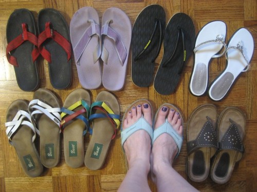 A variety of flip-flops and sandals arranged on a wooden floor, with a pair of feet in the center.