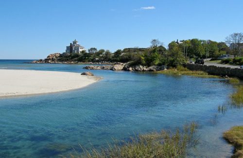 A serene coastal scene featuring a sandy beach, clear blue water, and a distant house surrounded by greenery.