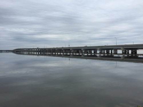 A bridge spans a calm body of water, reflecting the cloudy sky above.