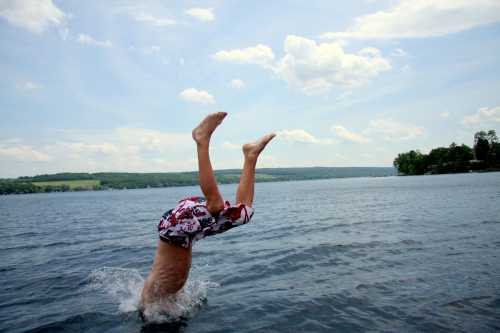 A child performs a handstand in the water, creating splashes against a backdrop of blue sky and green hills.