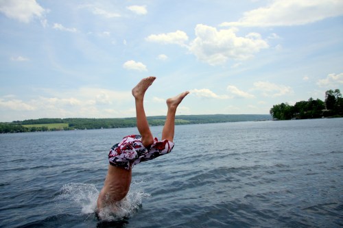 A child performs a handstand in the water, creating splashes against a backdrop of blue sky and green hills.