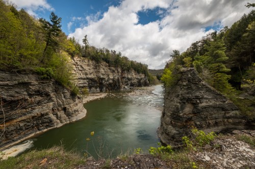 A serene river flows between rocky cliffs, surrounded by lush greenery and a partly cloudy sky.