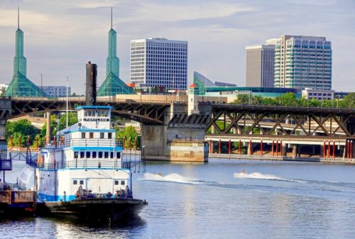 A riverboat on a calm river with a city skyline and bridges in the background under a clear sky.