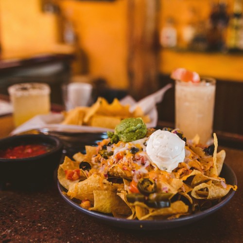 A plate of nachos topped with cheese, guacamole, sour cream, and olives, with drinks and tortilla chips in the background.