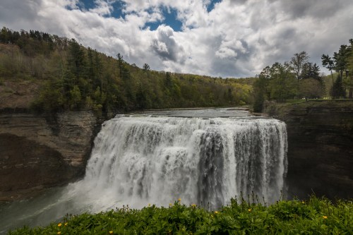 A majestic waterfall cascades over rocky cliffs, surrounded by lush greenery and a cloudy sky.
