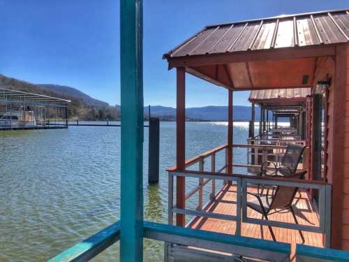 View of a serene lake with wooden cabins and chairs on a sunny day, surrounded by mountains in the background.