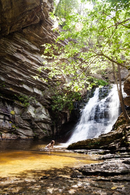 A serene waterfall cascades into a clear pool, surrounded by lush greenery and rocky cliffs. A person wades in the water.