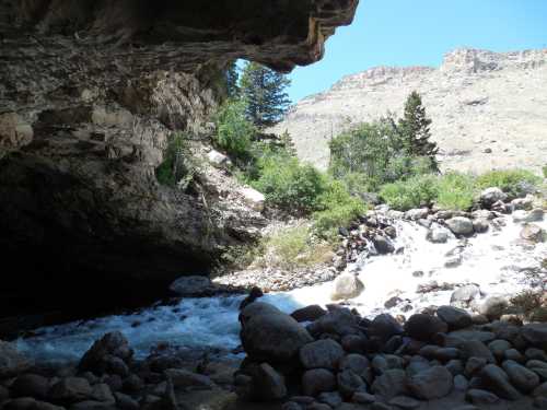 A rocky riverbank under a cave, with flowing water and trees against a backdrop of mountains and clear blue sky.