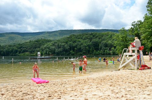 A sandy beach with people swimming and playing in the water, surrounded by trees and mountains under a cloudy sky.