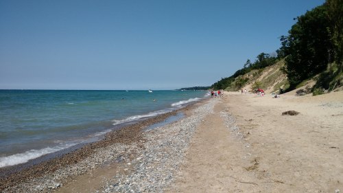 A sandy beach with pebbles, clear blue water, and people enjoying the sun under a clear sky. Trees line the shore.